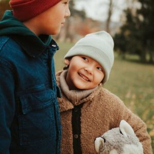 Two kids in warm jackets and hats enjoying the outdoors with a toy husky dog in autumn.