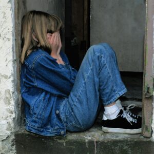 A young girl in denim sits alone in a doorway looking distressed, conveying loneliness.